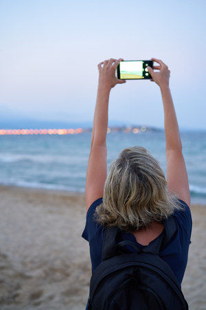 Woman taking a photo of a sea on smart phone on sunset at the beach, back view. Focus on womanの写真素材