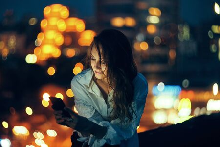 Young woman holding a smart phone in hands texting in the evening with city light on backgroundの写真素材