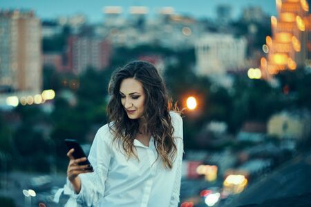 Portrait of young woman over office buildings holding a smart phone in a hand texting, summer duskの写真素材