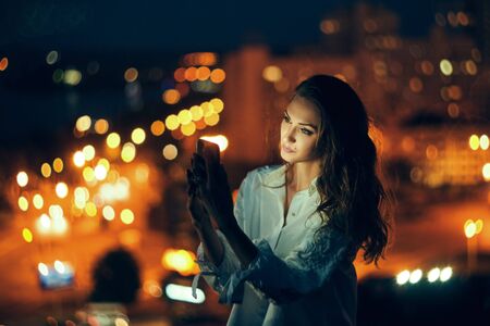 Young woman holding a smart phone in hands texting in the evening with city light on backgroundの写真素材