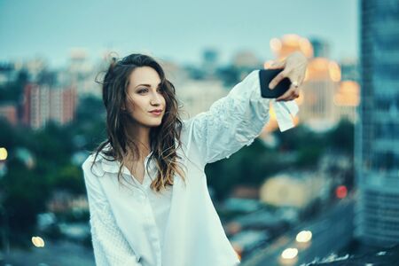 Young beautiful woman making self portrait with her smart phone over cityscape on dusk or video blogging, with evening lightings on backgroundの写真素材