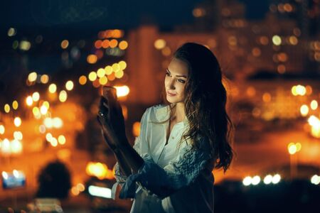 Young woman holding a smart phone in hands texting in the evening with city light on backgroundの写真素材