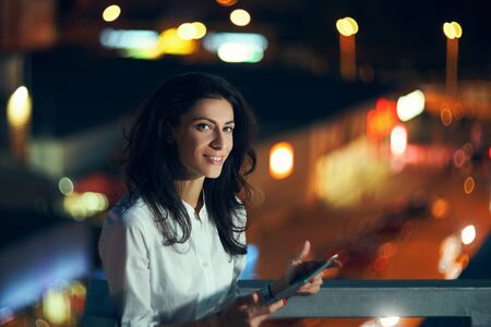 Young woman holding a digital tablet in hands texting at night with city lights on backgroundの写真素材