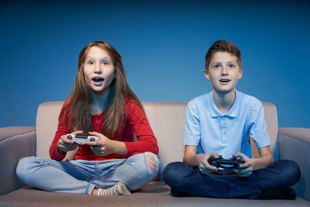 Computer game competition. Gaming concept. Excited children sitting on sofa playing video game with joysticksの写真素材