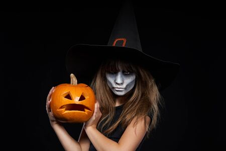 Serious Halloween witch holding Jack-O-Lantern pumpkin looking at camera on black background. Preteen girl with terrifying makeup and witch hat.の写真素材
