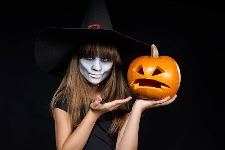 Halloween witch girl showing Jack-O-Lantern pumpkin on palm looking at camera on black background. Preteen girl with terrifying makeup and witch hat.の写真素材
