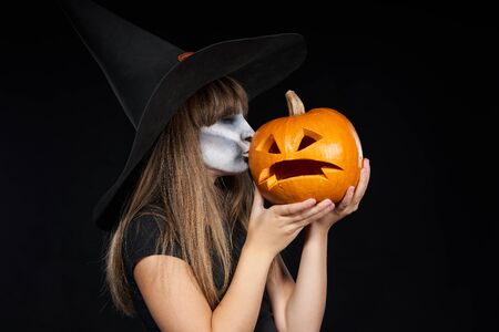 Halloween witch girl giving a kiss to Jack-O-Lantern pumpkin, on black background. Preteen girl with terrifying makeup and witch hat.の写真素材