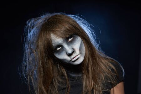 Halloween zombie. Closeup portrait of a girl with zombie makeup looking at camera over dark background with smoke and backlightの写真素材