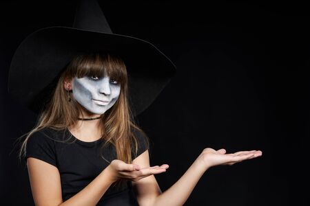 Halloween witch girl looking at camera showing blank copy space on her palms, on black background. Preteen girl with terrifying makeup and witch hat.の写真素材