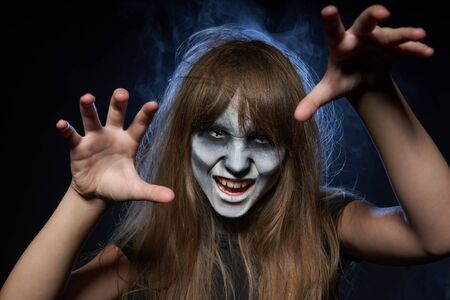 Halloween zombie. Closeup portrait of a girl with zombie makeup showing her teeth over dark background with smoke and backlightの写真素材