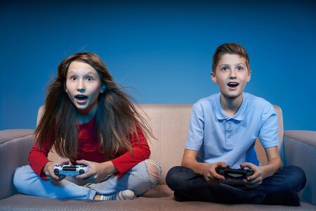 Computer game competition. Gaming concept. Excited children sitting on sofa playing video game with joysticksの写真素材