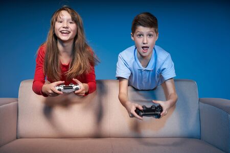 Computer game competition. Gaming concept. Excited children leaning on the sofa back playing video game with joysticksの写真素材