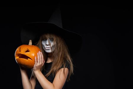 Serious Halloween witch holding Jack-O-Lantern pumpkin looking at camera on black background. Preteen girl with terrifying makeup and witch hat.の写真素材
