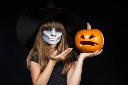 Halloween witch girl showing Jack-O-Lantern pumpkin on palm looking at camera on black background. Preteen girl with terrifying makeup and witch hat.の写真素材