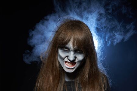 Halloween zombie. Closeup portrait of a girl with zombie makeup showing her teeth over dark background with smoke and backlightの写真素材
