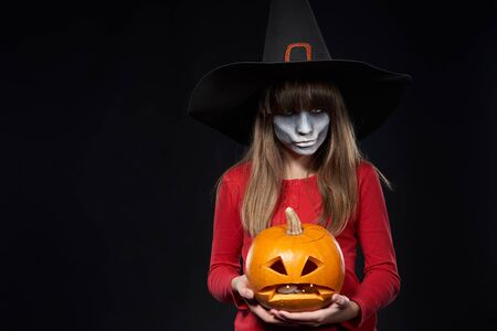 Serious Halloween witch holding Jack O'Lantern pumpkin looking at camera on black background. Preteen girl with terrifying makeup and witch hat.の写真素材
