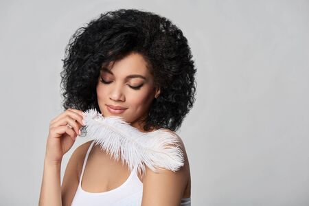 Closeup portrait of beautiful mixed race woman touching her skin with white ostrich feather enjoying its softness, studio shotの写真素材
