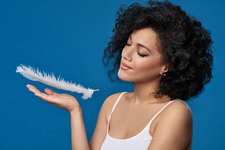 Closeup portrait of beautiful mixed race woman catching white ostrich feather falling on her palm enjoying its softness and beauty, studio shotの写真素材
