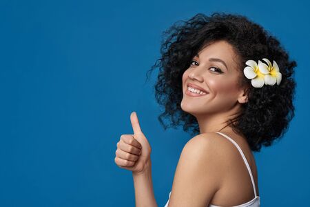 Closeup portrait of beautiful mixed race woman exotic plumeria flower in her hair looking at camera and gesturing thumb up, over blue backgroundの写真素材