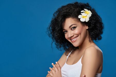 Closeup portrait of beautiful mixed race woman exotic plumeria flower in her hair looking at camera smiling, over blue backgroundの写真素材