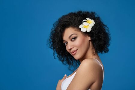 Closeup portrait of beautiful mixed race woman exotic plumeria flower in her hair looking at camera alluring, over blue backgroundの写真素材