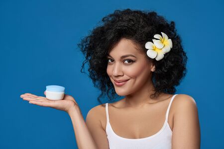 Closeup of beautiful mixed race woman exotic plumeria flower in her hair holding face cream on her palm, over blue backgroundの写真素材