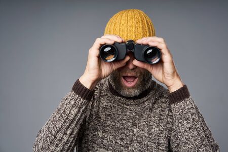 Excited surprised mature man in warm sweater and knitted hat looking at camera through binoculars, over grey studio backgroundの写真素材