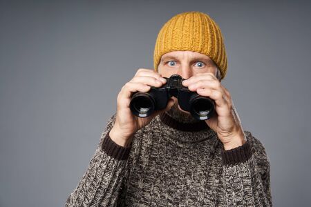 Shocked mature man in warm sweater and knitted hat looking at camera holding binoculars by his eyes, focus on binoculars, over grey studio backgroundの写真素材