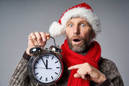 Closeup of mature man wearing Santa hat holding big alarm clock time approaching midnight, looking...