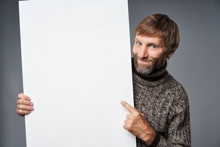 Smiling mature man in warm sweater holding white banner and pointing at blank copy space at banner, over grey studio backgroundの写真素材