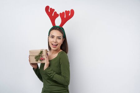 Happy girl wearing Christmas deer horns holding a gift box wrapped in craft paper with a branch of fir tree, looking at cameraの写真素材