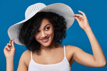 Closeup portrait of playful mixed race woman wearing summer straw hat with wide brims, looking to side over blue backgroundの写真素材
