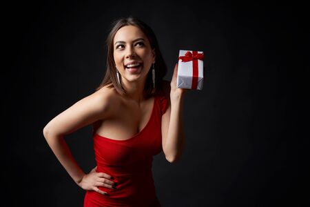 Happy playful woman in red evening gown holding a gift box, looking away, over black backgroundの写真素材