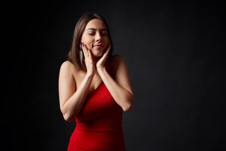 Excited surprised woman in red evening gown with hands on cheeks dreaming with closed eyes, over black backgroundの写真素材