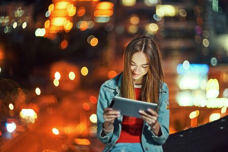 Lovely teen girl on night cityscape background with street lights, using a digital tabletの写真素材