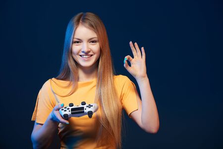 Gamer girl. Happy girl holding video game joystick showing OK sign, on blue backgroundの写真素材
