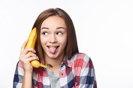 Closeup portrait of a teen girl holding banana like a phone, looking to side with funny expression showing her tongueの写真素材
