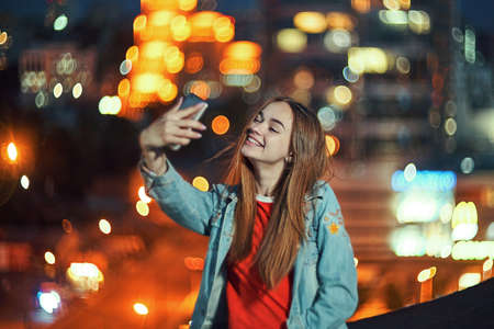 Lovely teen girl on cityscape background making self portrait with her smart phone, evening street light bokeh on backgroundの写真素材