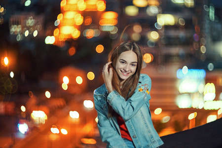 Lovely teen girl on night cityscape background with street lights, smiling at cameraの写真素材