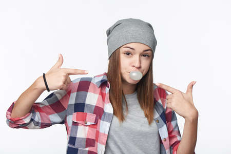 Closeup of cheeky teen girl blowing bubblegum and pointing at it, looking at camera, studio portraitの写真素材