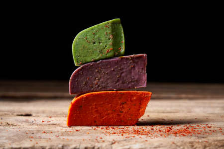 Three different gourmet cheeses in pile on rough wooden planks: lavender, paprika and pesto cheeses. over dark background, shallow depth of fieldの写真素材