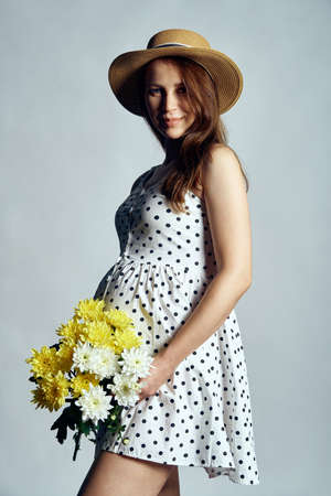 Pregnant woman in summer dress and straw hat holding bunch of flowers, studio portrait, isolatedの写真素材