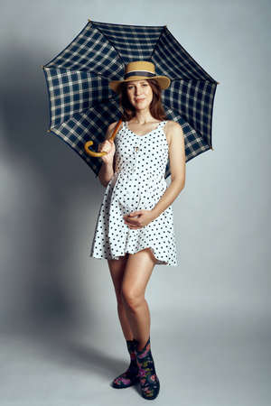 Pregnant woman in country style summer dress and straw hat standing under umbrella, studio portraitの写真素材