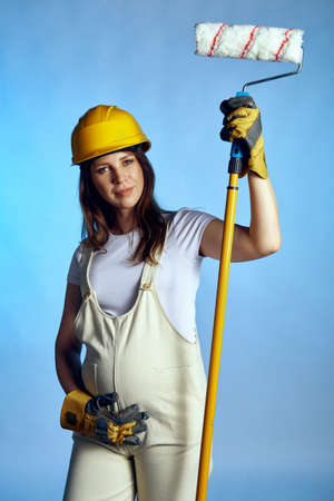 Pregnant woman wearing builder overall and protective helmet holding painting rollerの写真素材