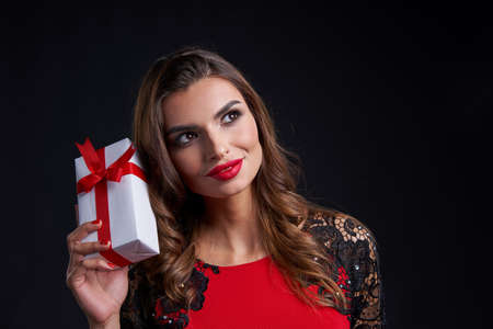 Holidays and celebration concept. Closeup of thinking gorgeous woman with red lipstick makeup holding gift box with red ribbon, looking to side at blank copy space, over dark backgroundの写真素材