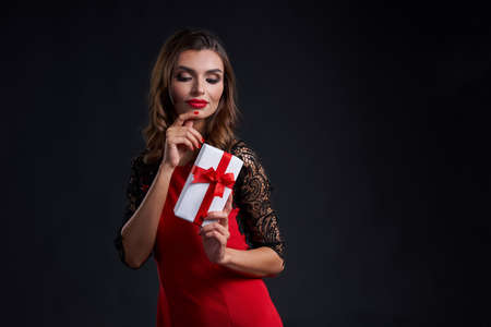 Holidays and celebration concept. Thinking gorgeous woman with red lipstick makeup holding gift box with red ribbon, over dark backgroundの写真素材
