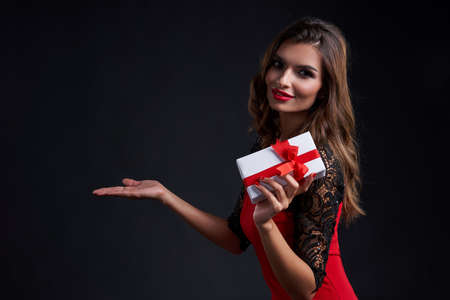 Holidays and celebration concept. Smiling gorgeous woman with red lipstick makeup holding gift box with red ribbon, holding blank space on open palm, over dark backgroundの写真素材