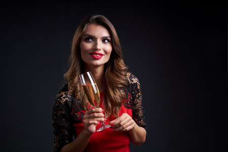 Holidays and celebration concept. Smiling gorgeous woman with red lipstick makeup holding a glass with champagne, over dark backgroundの写真素材