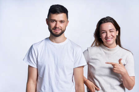 Selecting him. Young smiling couple standing over light background, joyful female pointing at guy and winkingの写真素材