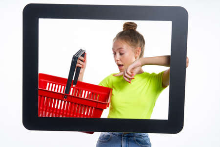 Online shopping concept. Trendy teen girl looking through digital screen holding empty shopping basket, pointing at it with surprise, isolated on light backgroundの写真素材